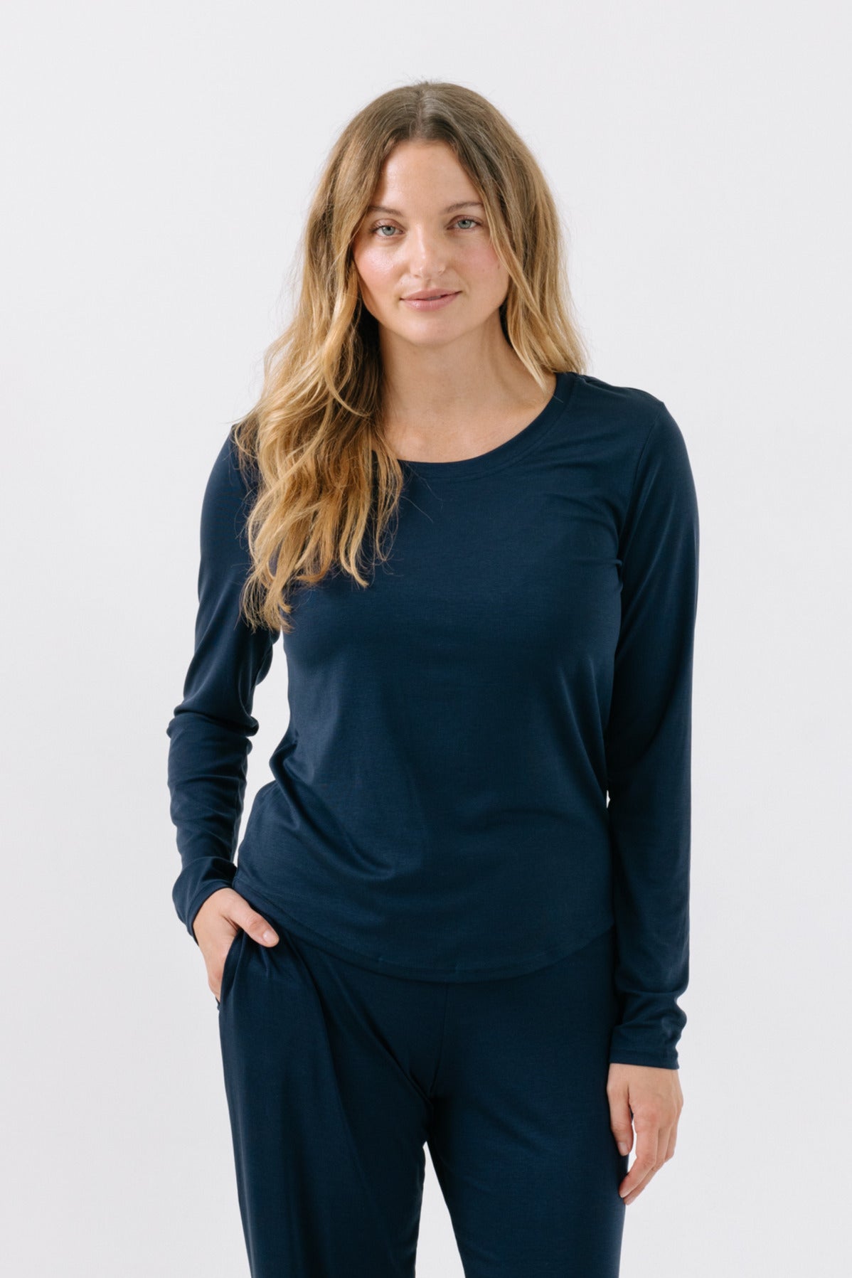 A woman wearing a navy, long-sleeve pajama t-shirt with her hand in her pocket on a white studio background.