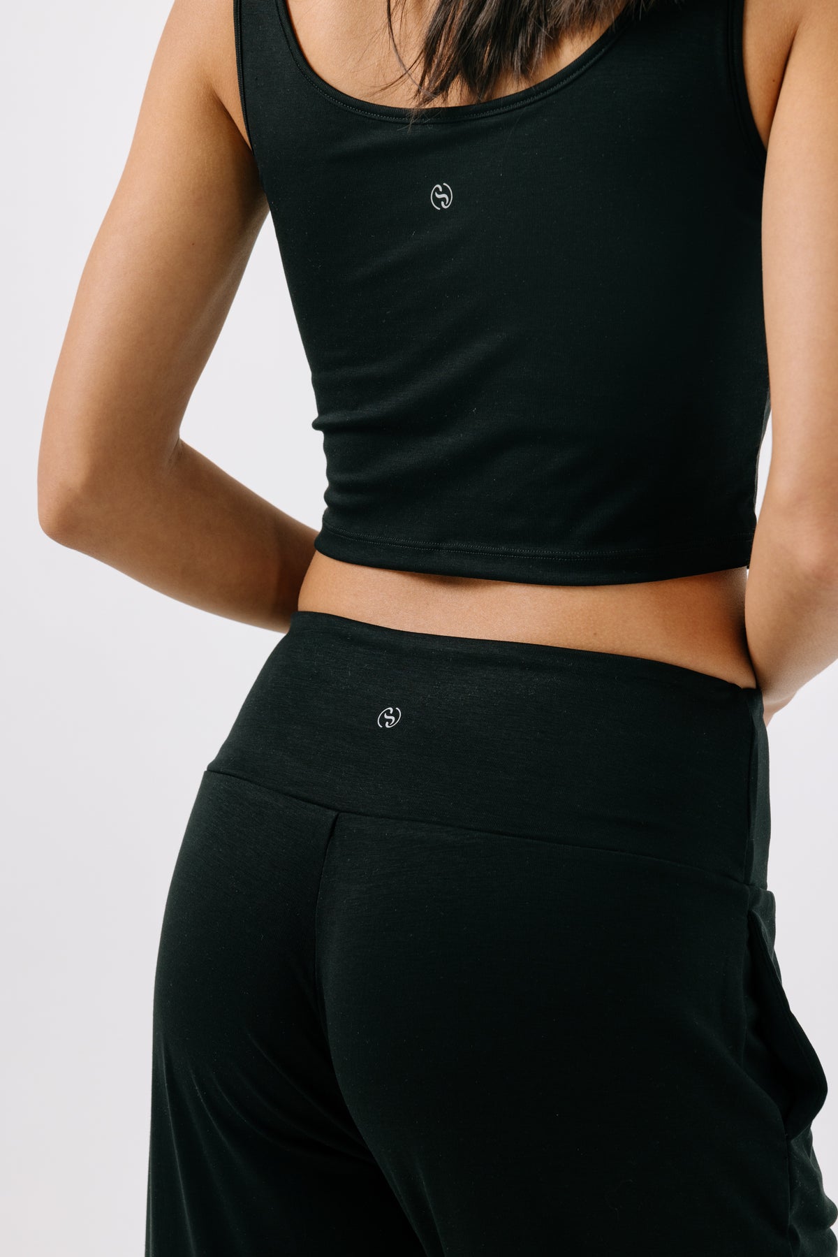Woman facing away from the camera wearing a black pajama crop top and matching black pajama pants on a white studio background.