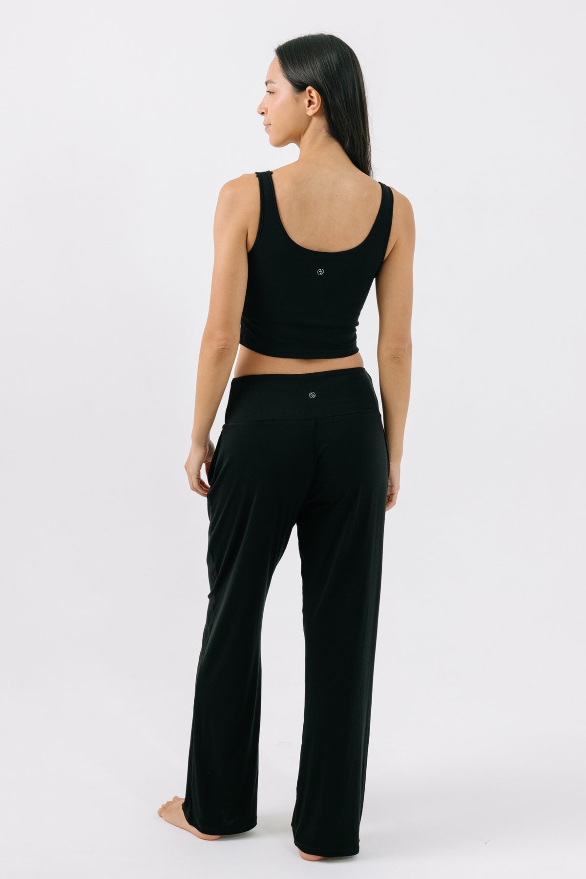 Woman facing away from the camera wearing a black pajama crop top and matching black pajama pants on a white studio background.