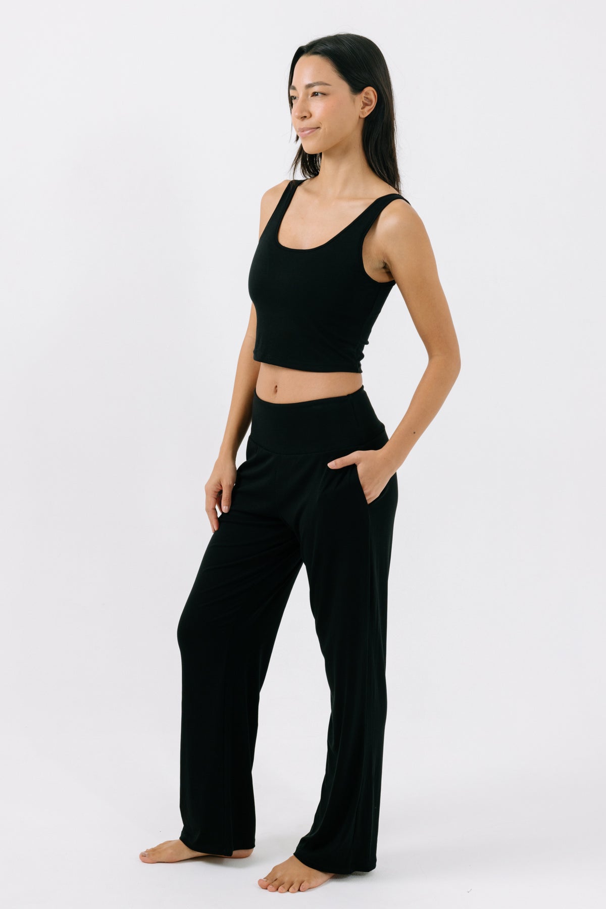 Woman wearing a black pajama crop top and matching black pajama pants with her hand in her pocket on a white studio background.