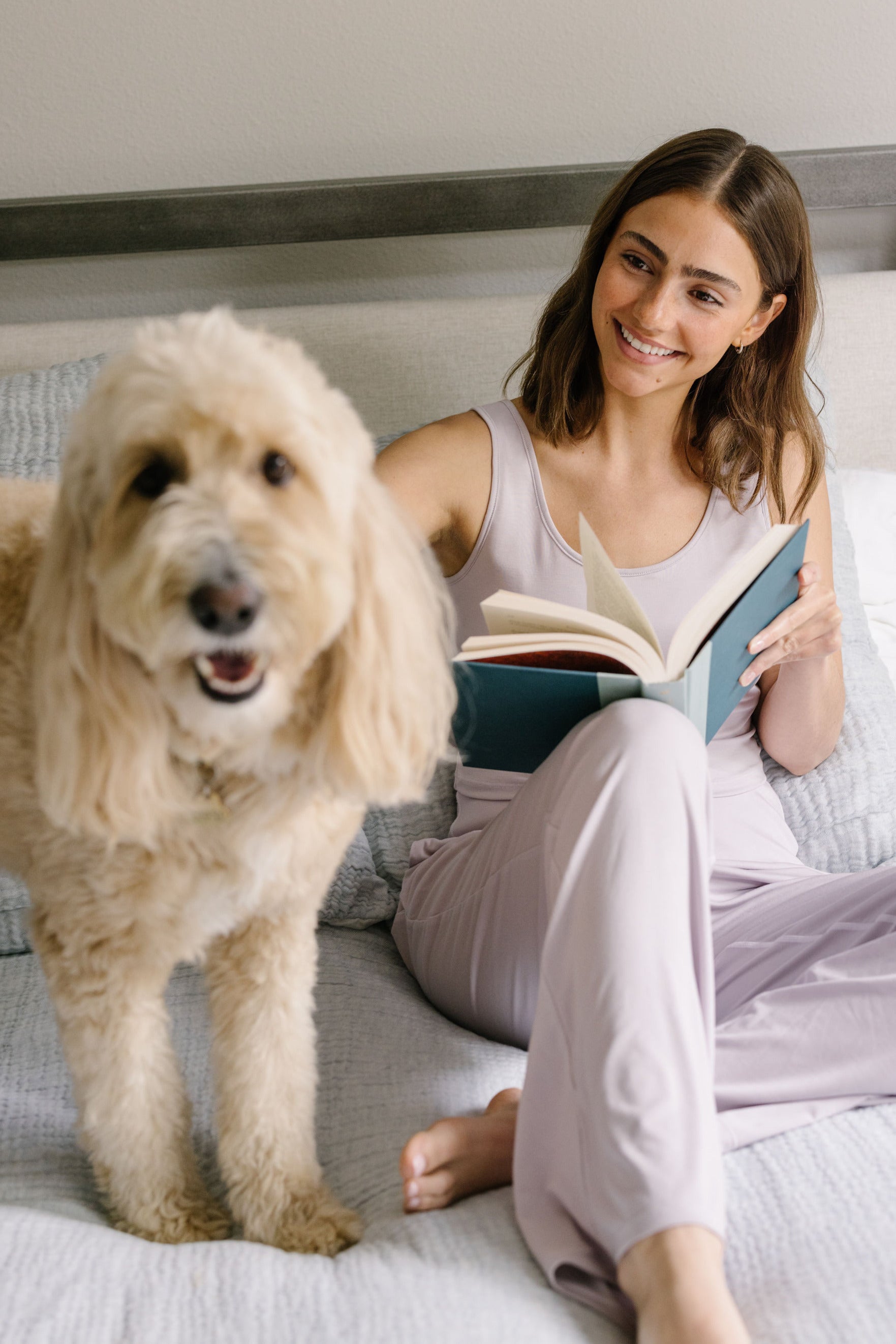 Woman reading a book on a bed wearing lavender pajamas with a dog next to her.