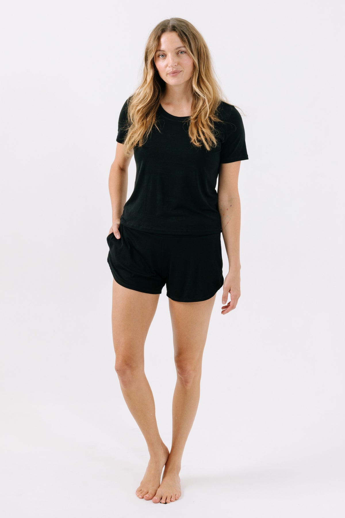 Woman wearing a black pajama t-shirt and matching black pajama shorts with her hand in her pocket on a white studio background.