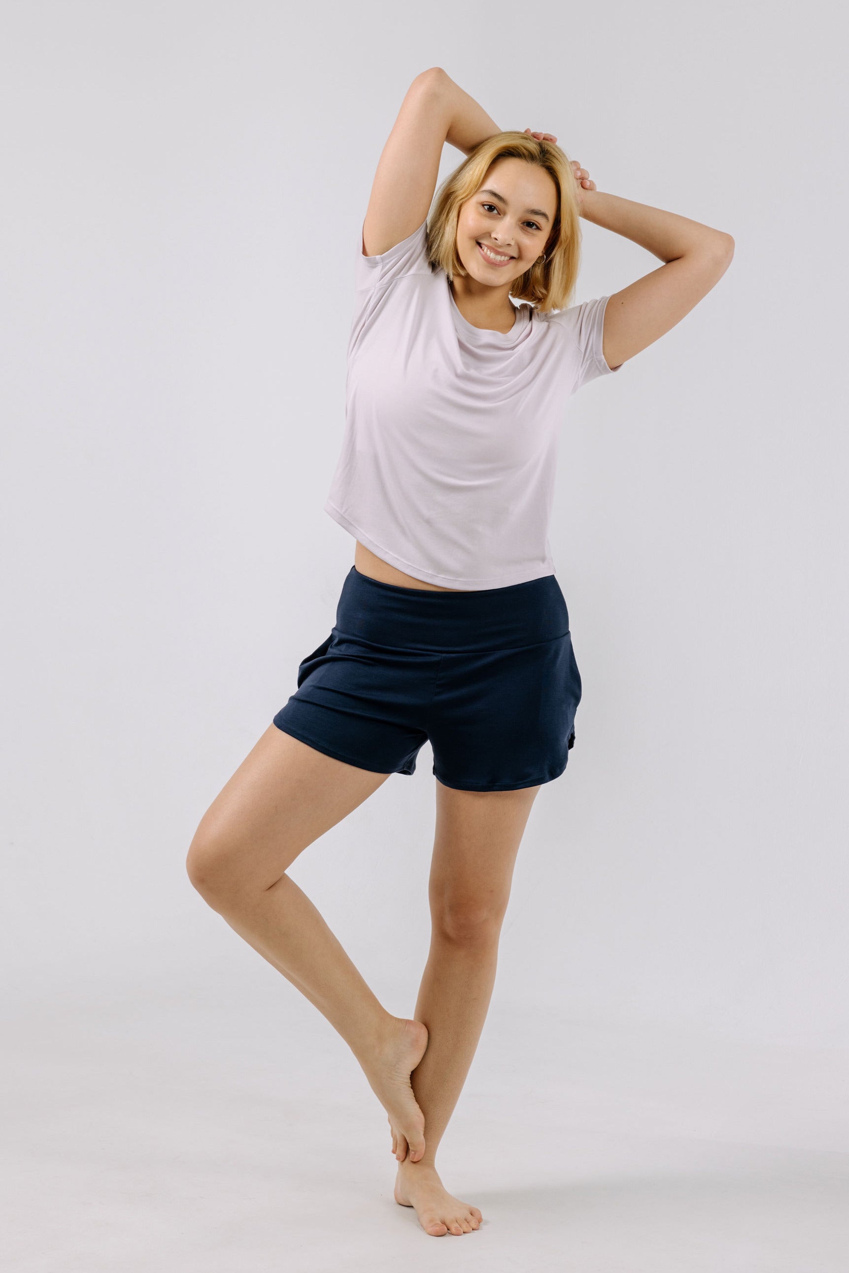 Woman posing with arms raised wearing a light purple pajama t-shirt and navy shorts on a white background.