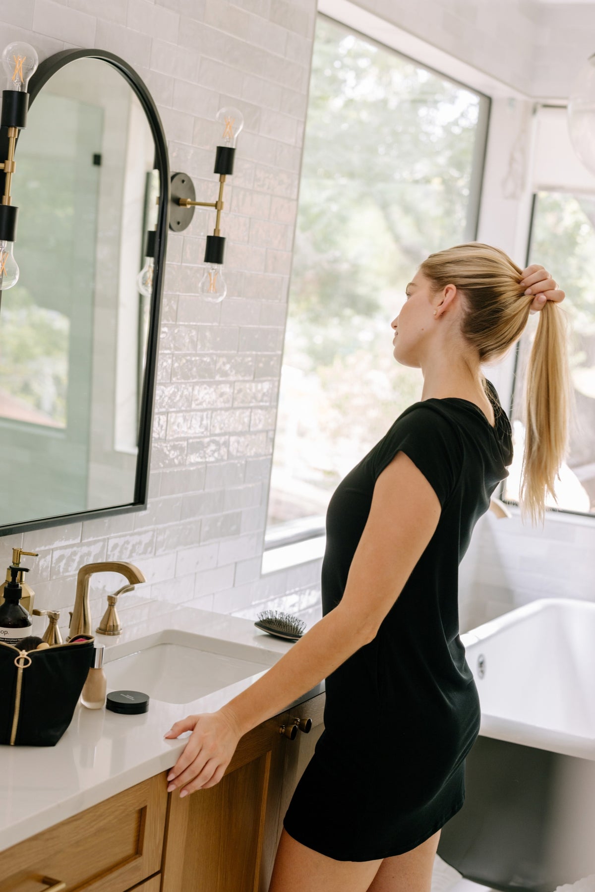A woman with blonde hair tied back in a ponytail stands in profile in a bright bathroom, wearing a short black pajama dress. She is positioned next to a white sink with a gold faucet, a large arched mirror, and a window looking out onto greenery.