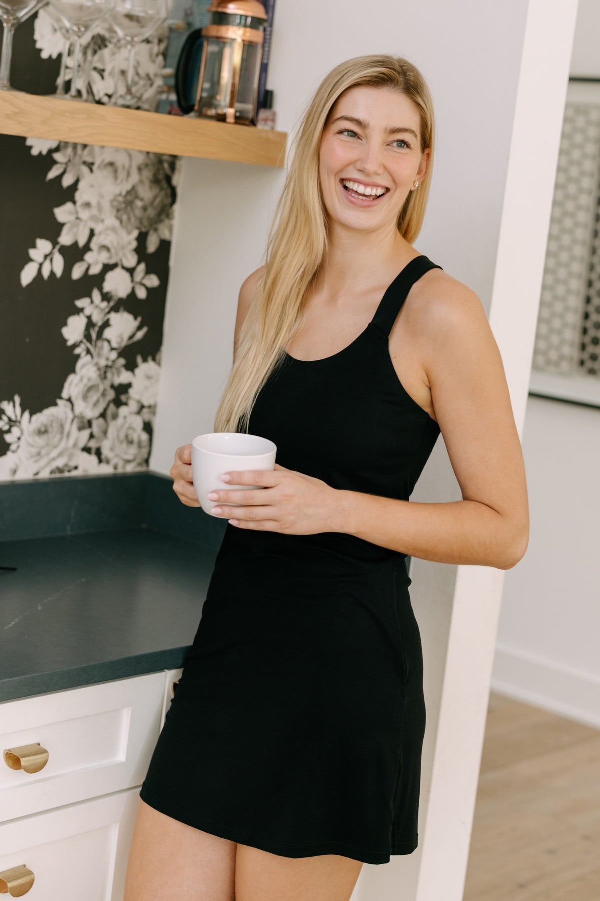 A woman wearing a black racerback pajama dress holding a coffee cup next to a counter in a home.