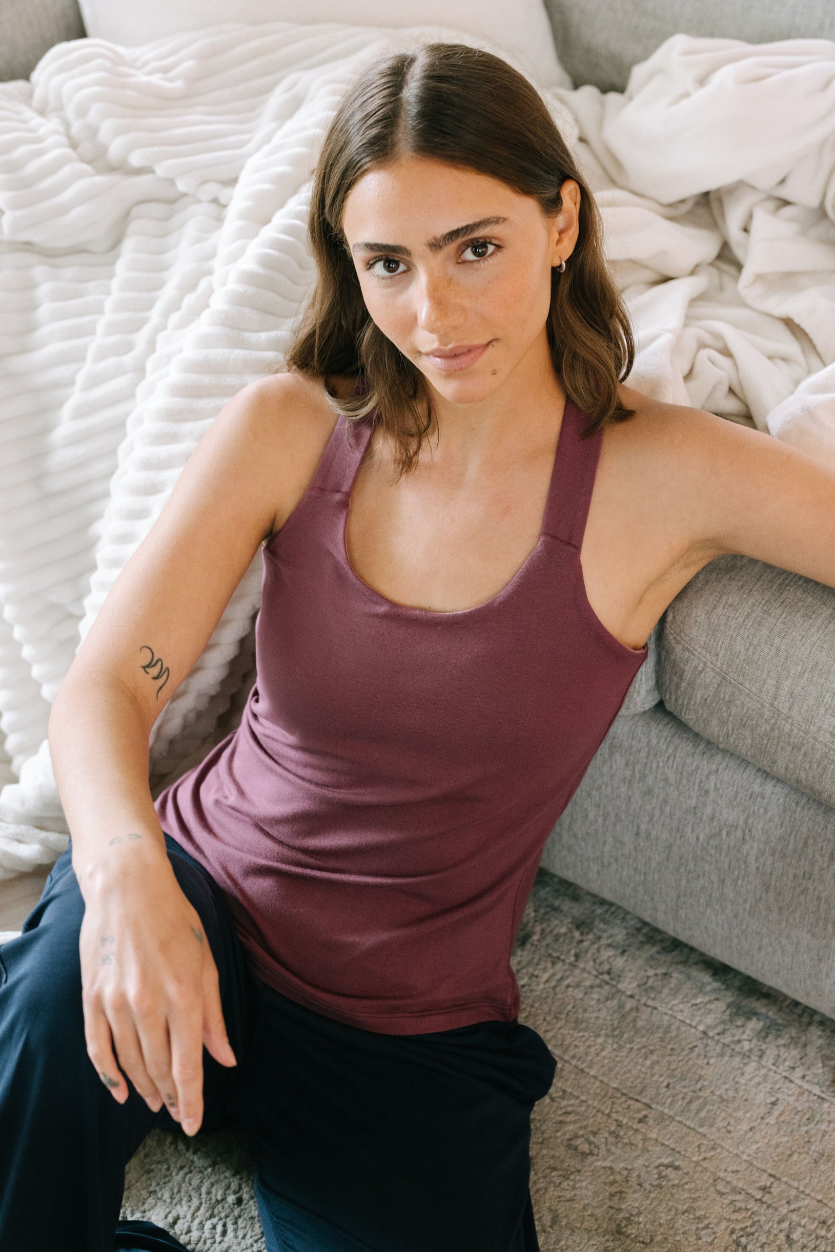 Woman in a plum pajama tank top leaning up against a couch with white pillows and blankets.