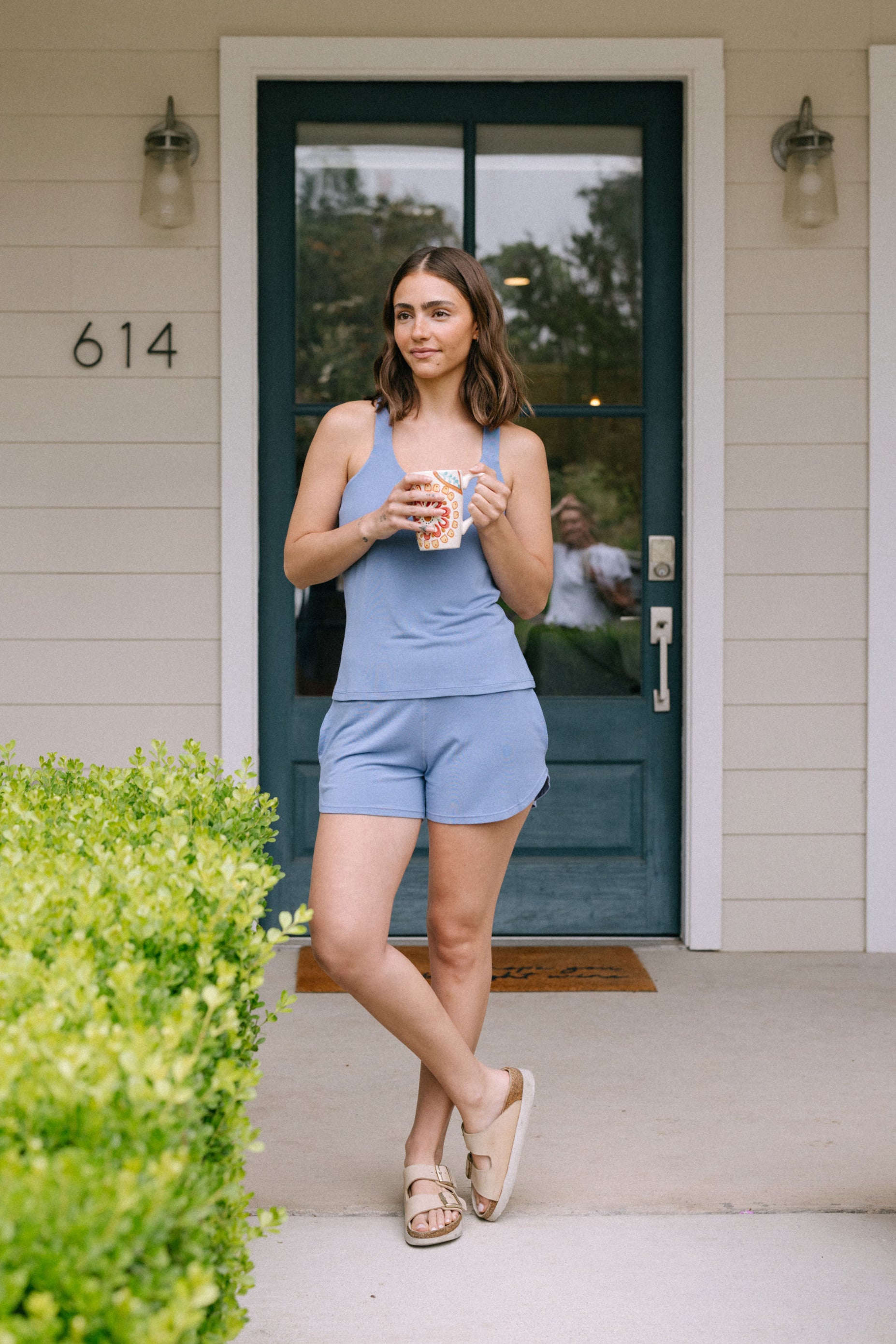 Woman holding a coffee cup on a porch wearing a periwinkle tank and shorts pajama set.