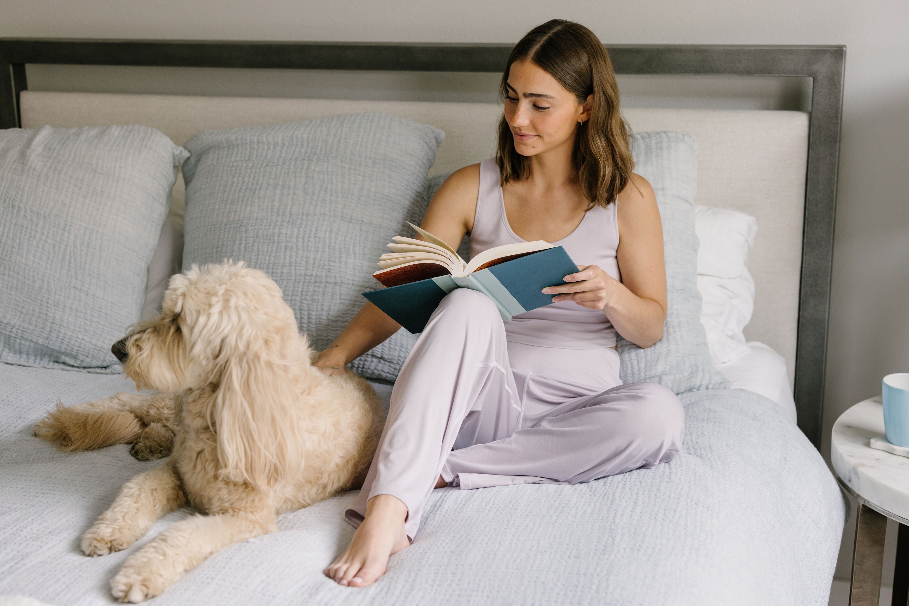 Woman reading a book on a bed with a dog while wearing lavender pajamas.