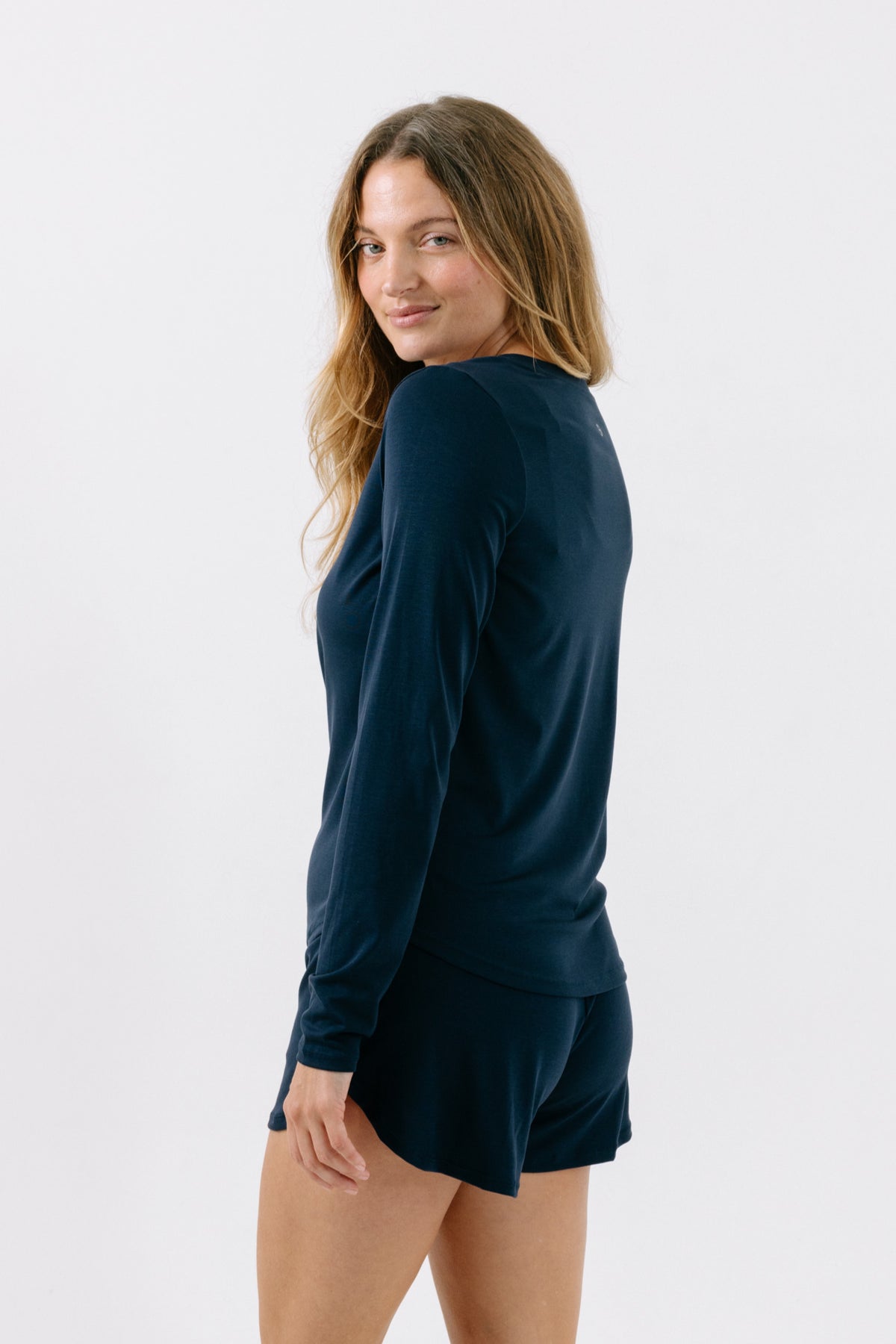 A woman showing the side and back of a navy, long-sleeve pajama t-shirt and matching shorts on a white studio background. 