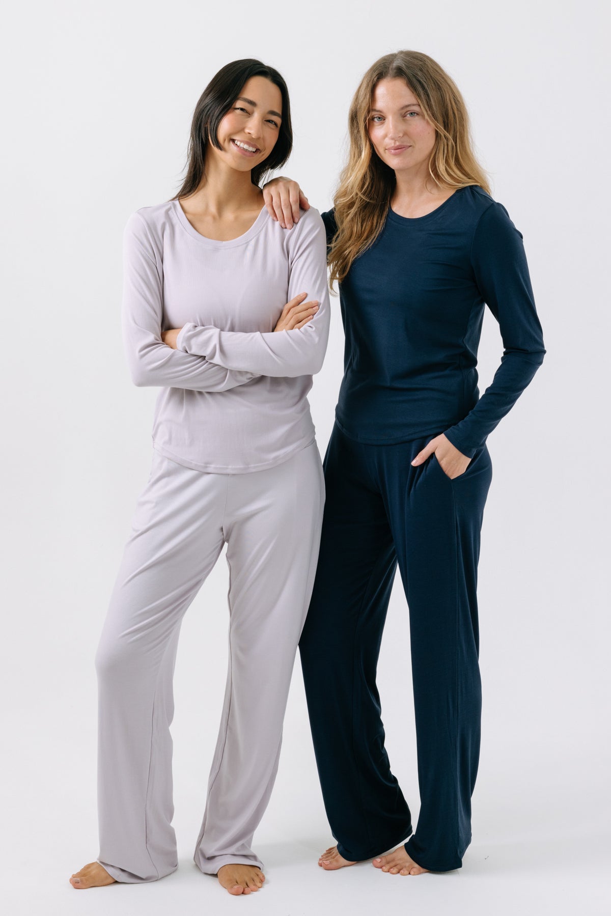 Two women wearing matching long-sleeve pajama t-shirts and pajama pants on a white studio background. One woman is wearing a lavender set. The other woman is wearing a navy set.