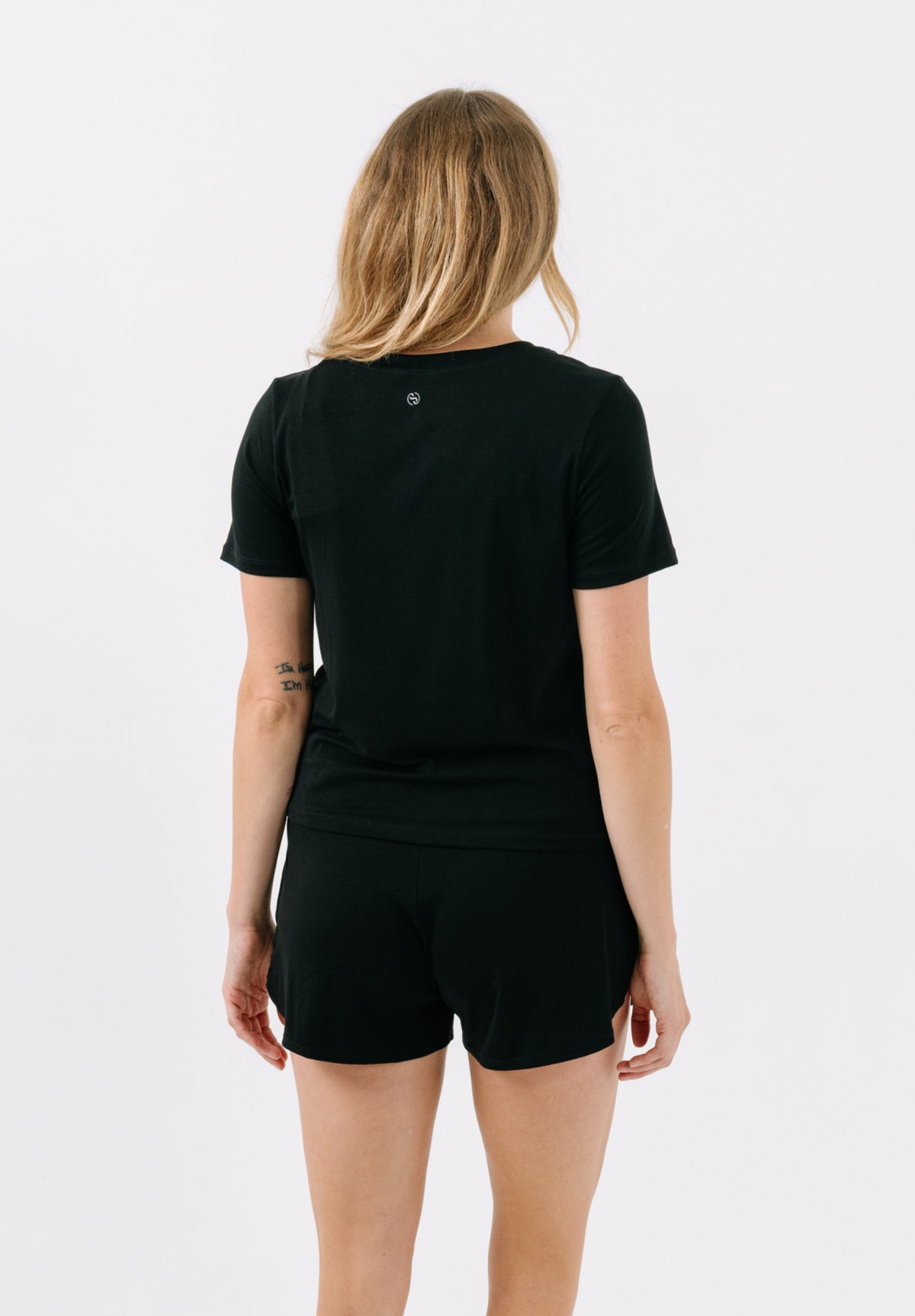 Woman facing away from the camera wearing a black pajama t-shirt and matching black pajama shorts on a white studio background.