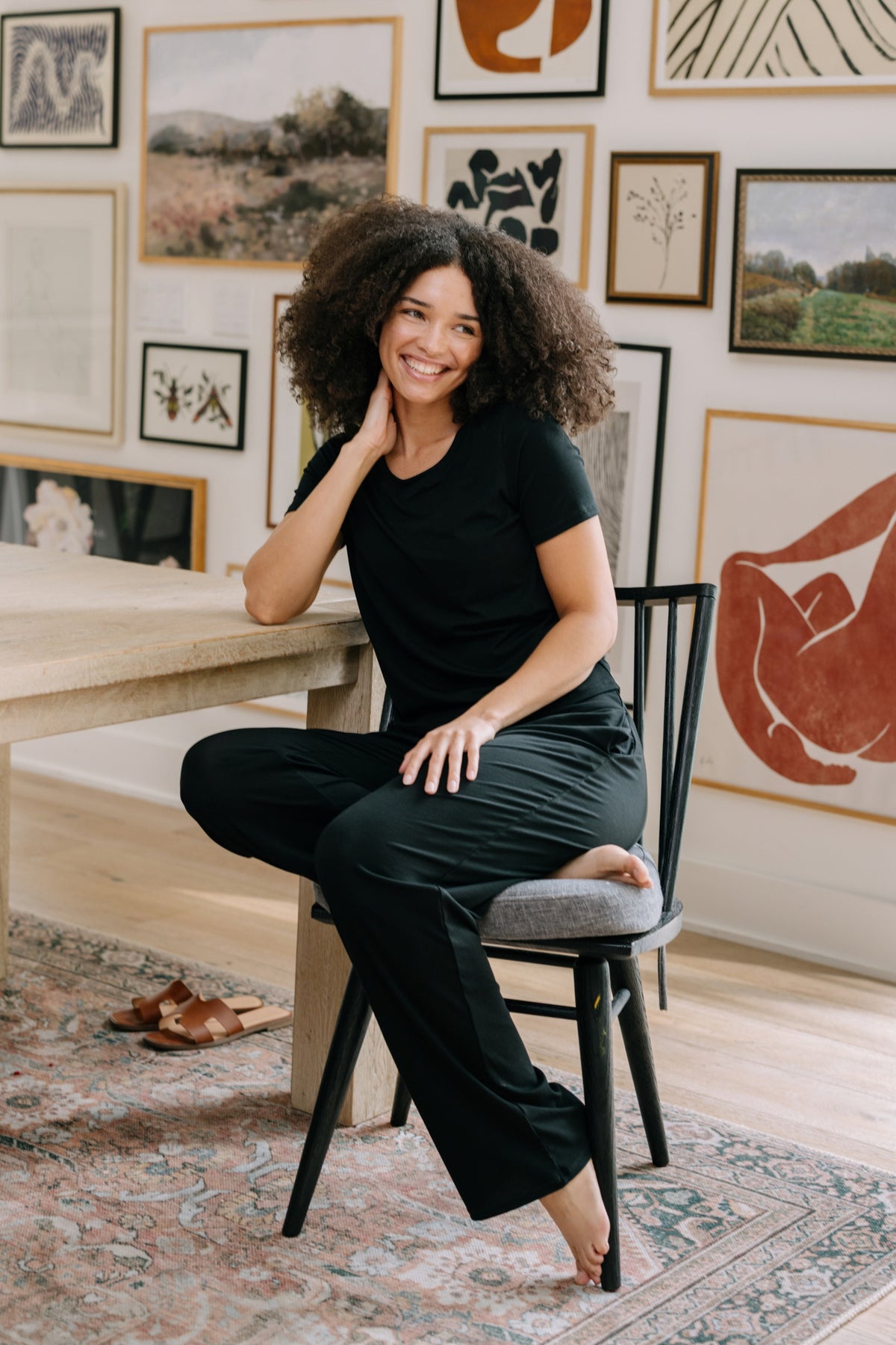 Woman sitting on a chair with artwork on the wall behind her wearing a black pajama t-shirt and matching black pajama pants. 