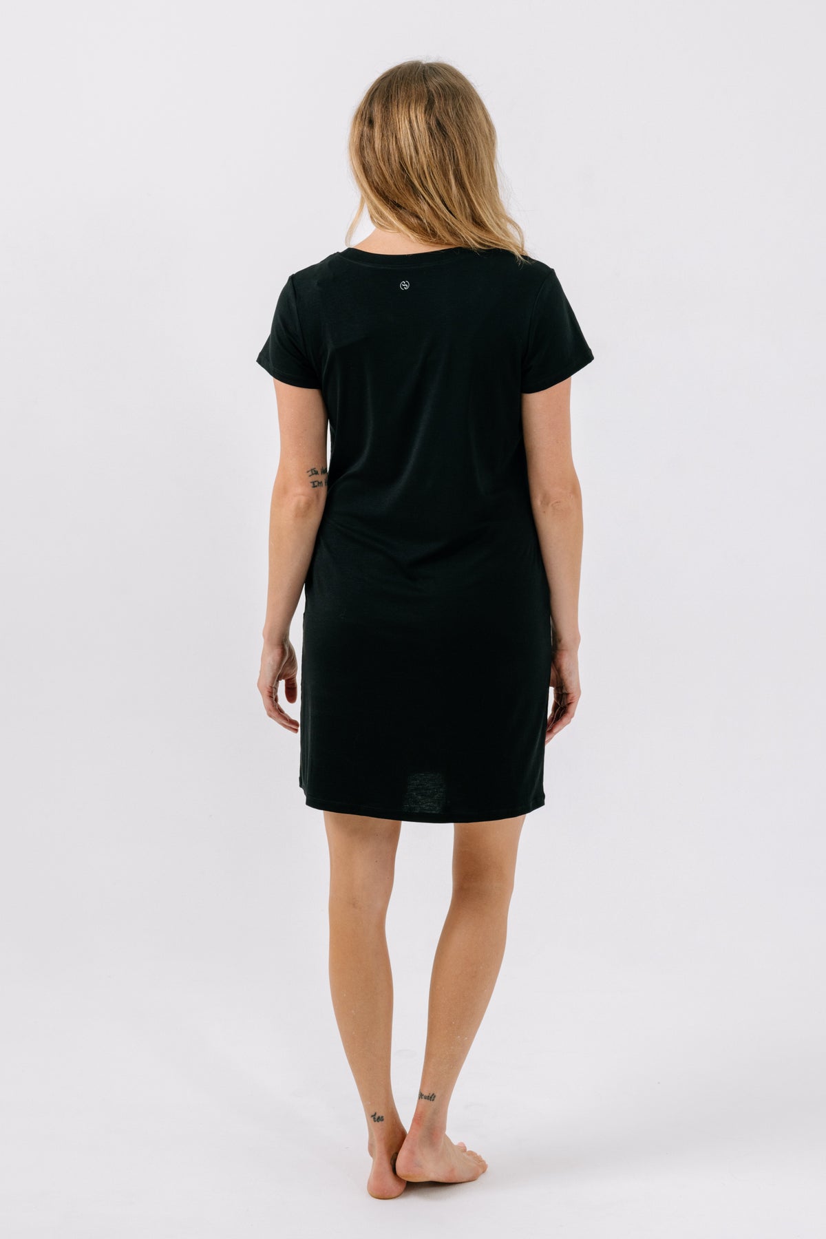 A woman viewed from behind wearing a black short-sleeve t-shirt pajama dress, standing barefoot against a plain white studio background.
