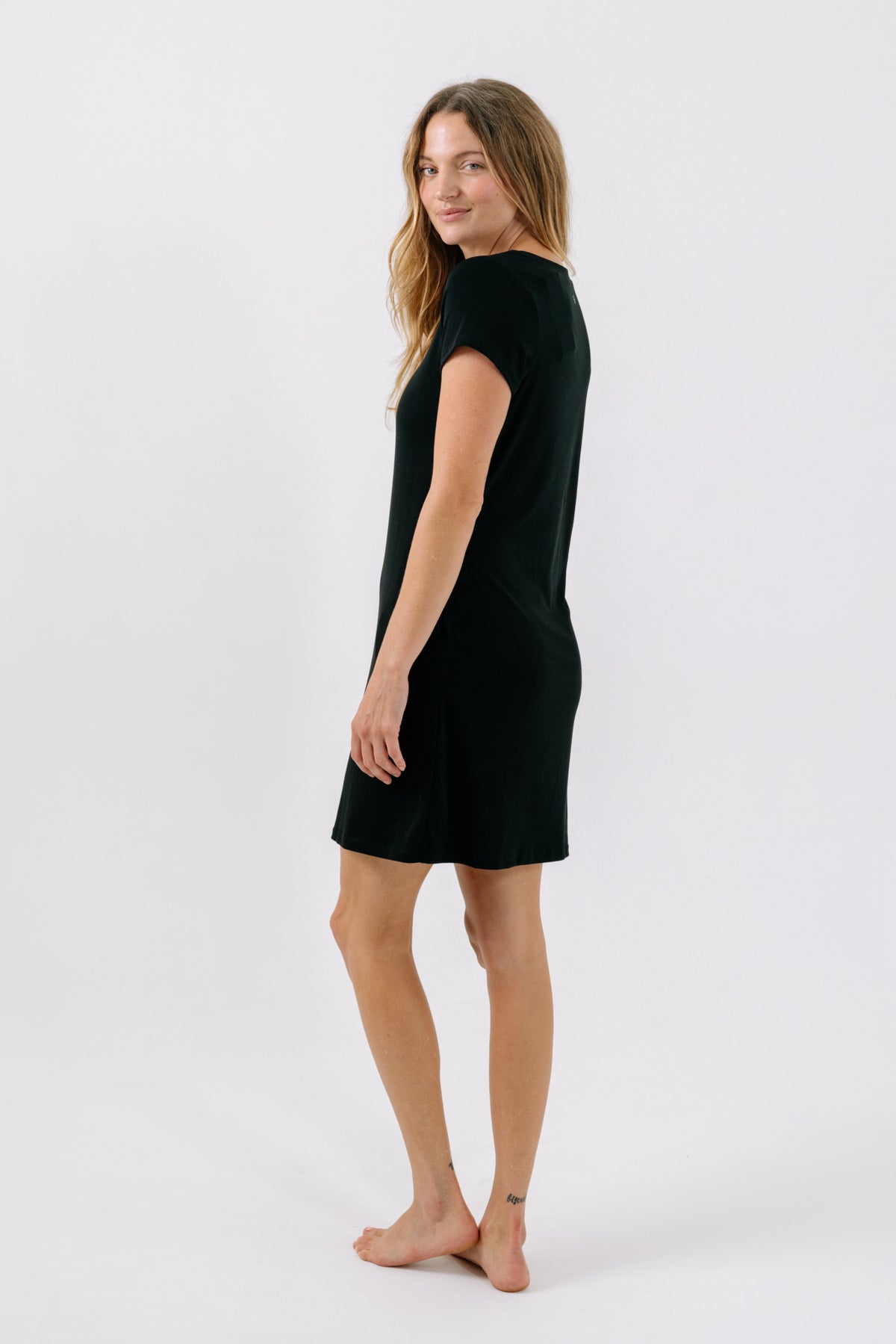 A woman modeling a short-sleeve black pajama dress, shown from a side-back angle against a plain white studio background.
