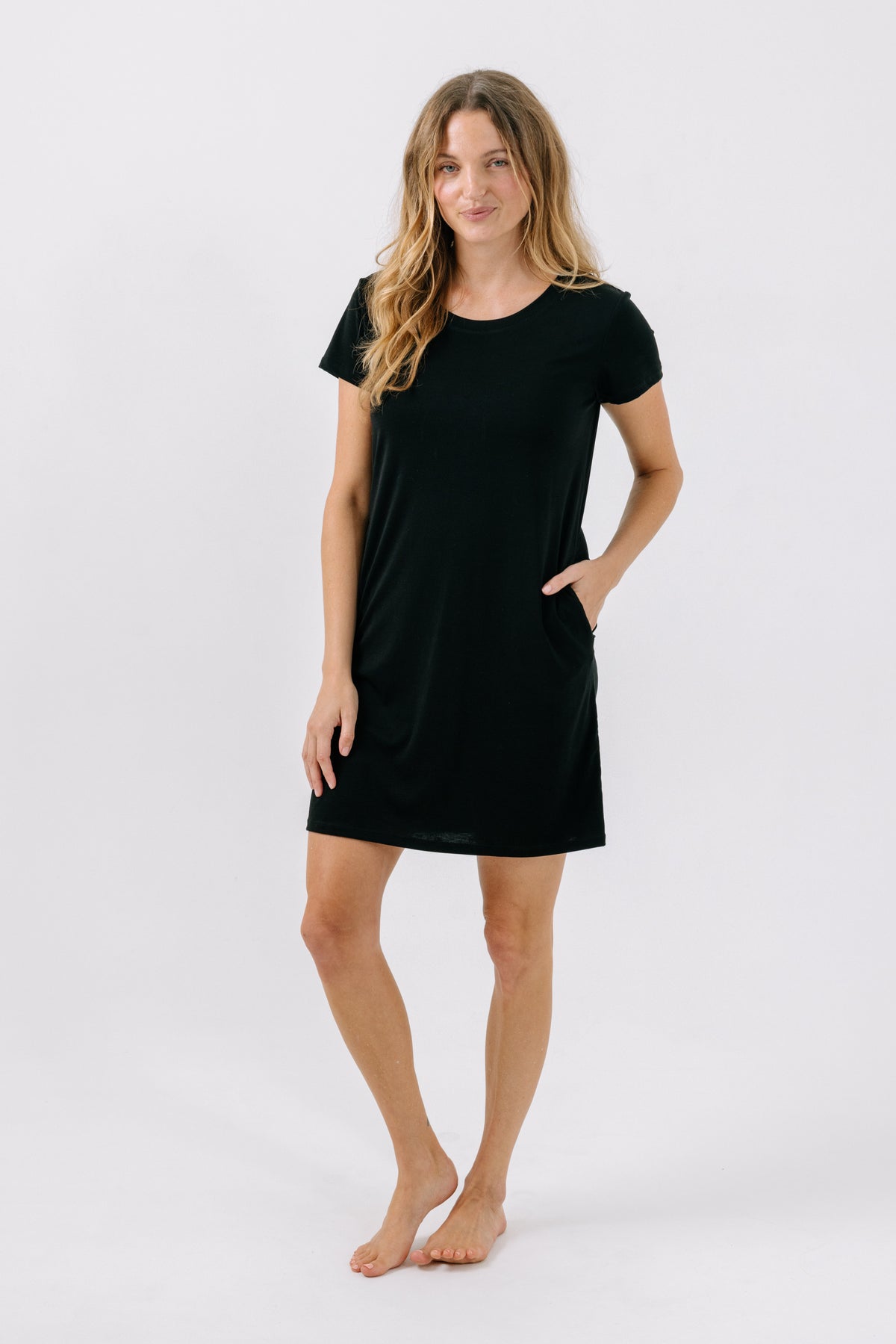 Woman wearing a black pajama t-shirt dress with her hand in her pocket on a white background.