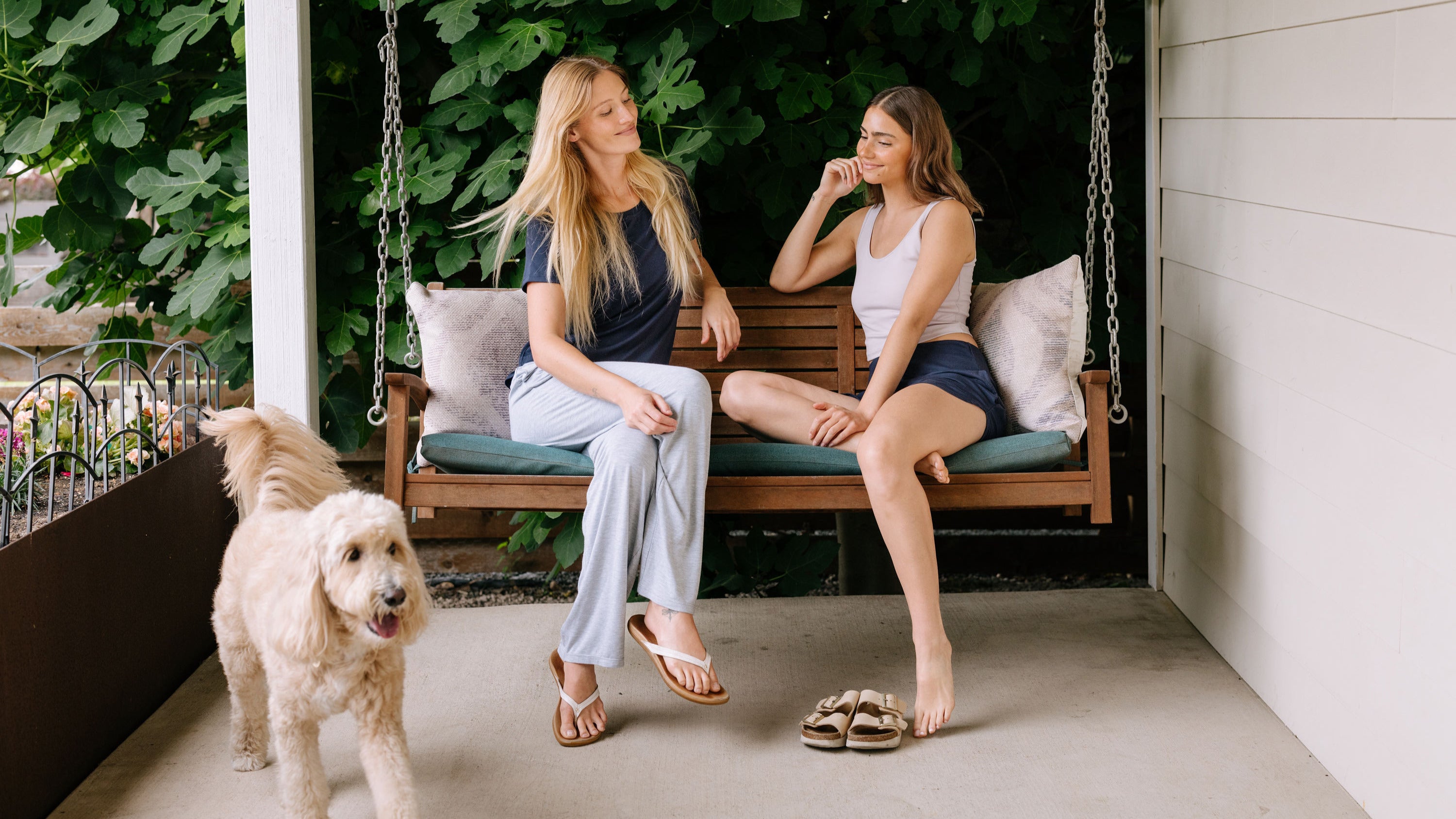 Two women sitting on a swing wearing pajamas with a dog standing nearby on a porch.
