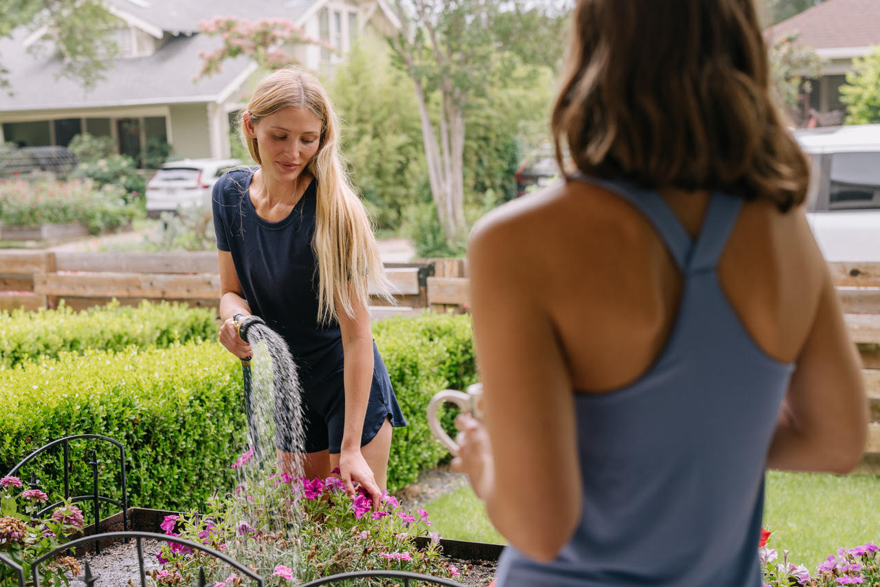 Two women in a garden, one watering plants and the other holding a cup.