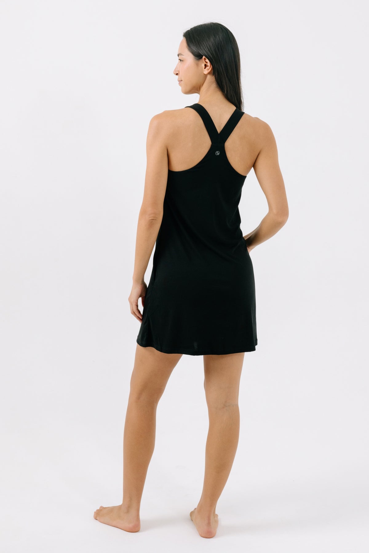 Woman wearing a black racerback pajama dress facing away from the camera on a white background.